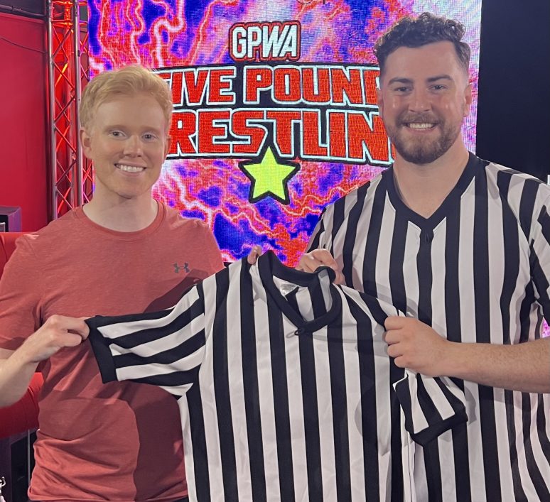 Carson Black graduades from the Professional Wrestling Referee Training Programme. He is holding one end of a striped referee shirt whilst Thomas Kearins holds the other. The "5 Pound Wrestling" logo, which is the show Carson debuted on, is displayed on the entrance screen in the background.